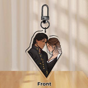 May include: A heart-shaped acrylic keychain featuring a couple, a man with dark hair and a woman with long brown hair, standing close together. The man is wearing a black jacket with gold buttons and the woman is wearing a black dress. The keychain has a silver metal ring.