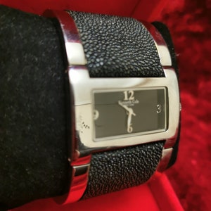 May include: A silver wristwatch with a black face and black leather band. The watch face has silver hands and Roman numeral markings. The band has a textured, pebbled design.