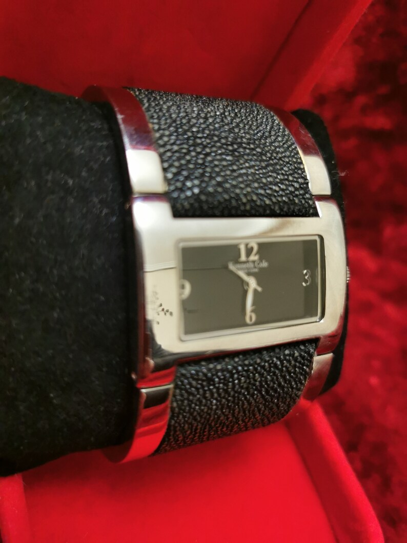 May include: A silver wristwatch with a black face and black leather band. The watch face has silver hands and Roman numeral markings. The band has a textured, pebbled design.