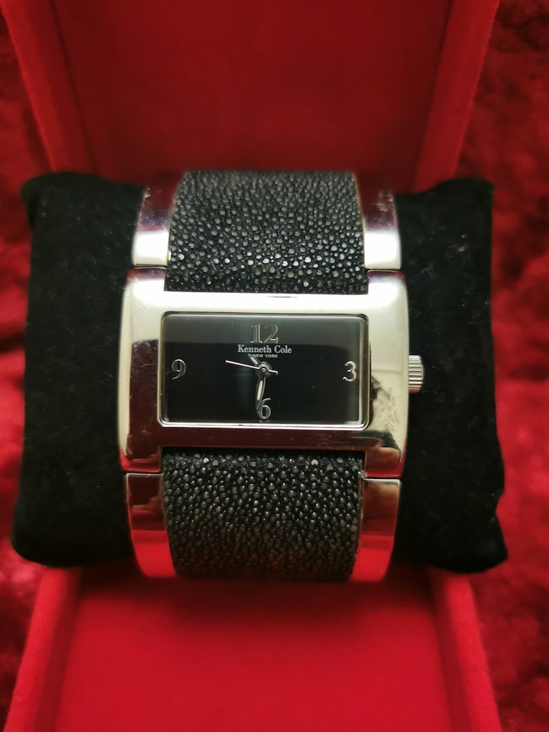 May include: A silver Kenneth Cole New York watch with a black face and black textured band.