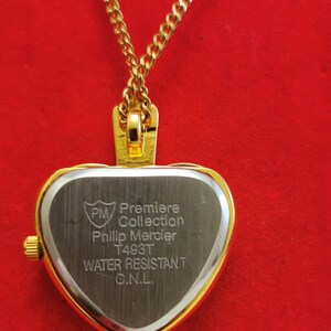 Op de afbeelding: Een goudkleurige hartvormige hangerhorloge aan een gouden ketting. De wijzerplaat is zilverkleurig met de tekst "PM Premiere Collection Philip Mercier T493T WATER RESISTANT C.N.L.". De achtergrond is effen rood.