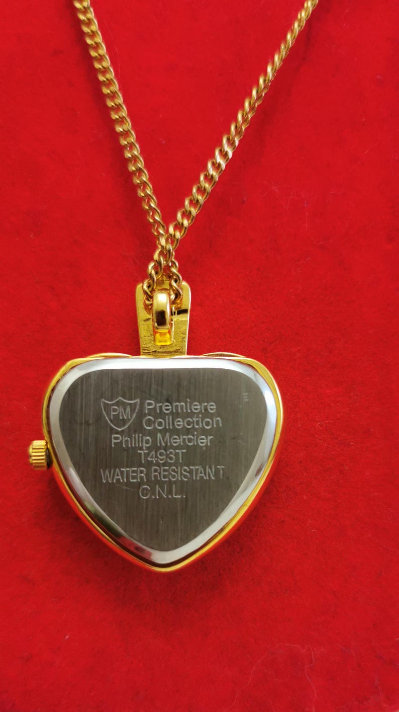 Op de afbeelding: Een goudkleurige hartvormige hangerhorloge aan een gouden ketting. De wijzerplaat is zilverkleurig met de tekst "PM Premiere Collection Philip Mercier T493T WATER RESISTANT C.N.L.". De achtergrond is effen rood.