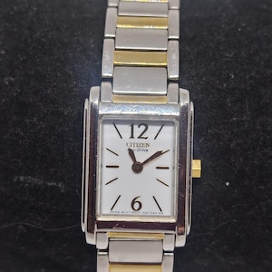May include: Citizen Eco-Drive watch with a rectangular white face, silver and gold-tone band. The watch face displays the Citizen logo, the numbers 12 and 6, and the hands. The band is made of silver and gold-tone metal links.