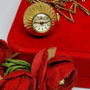 Pode incluir: Um relógio de bolso redondo dourado com um mostrador branco e ponteiros pretos. O relógio está em uma corrente dourada e está em uma caixa de veludo vermelho. O mostrador do relógio diz "17 Jewels" e "Bucherer".