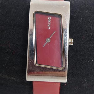 Könnte beinhalten: DKNY Armbanduhr mit rechteckigem, rotem Zifferblatt und silberfarbenem Metallgehäuse. Die Uhr hat ein rotes Lederarmband. Auf dem Zifferblatt ist das DKNY-Logo zu sehen. Das Armband ist ca. 18 cm lang.