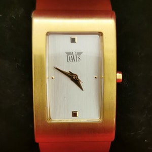 Può includere: Un orologio da polso dorato con un quadrante rettangolare. Il quadrante è bianco con segni neri e la parola "DAVIS" in lettere nere.
