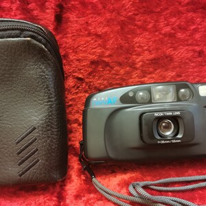 Op de afbeelding: Een zwarte en zilveren compactcamera met een Ricoh Twin Lens, een 3-modus flitser en een zwarte leren draagtas met een band.