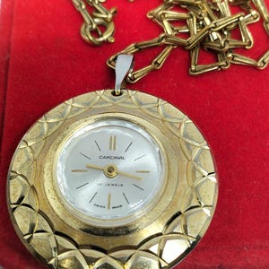 Op de afbeelding: Een goudkleurig hangerhorloge met een gouden ketting. De wijzerplaat is wit met zwarte wijzers en het woord "Cardinal". Het horloge is gemarkeerd met "17 Jewels" en "Swiss Made". Het horloge ligt op een rode ondergrond.