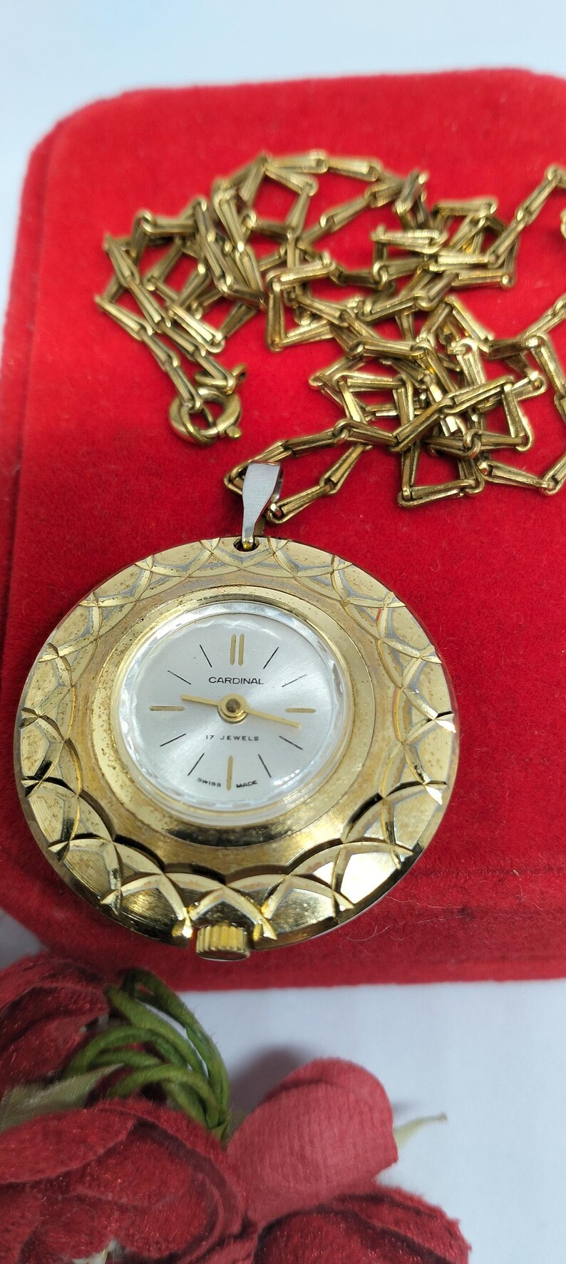 Op de afbeelding: Een goudkleurig hangerhorloge met een gouden ketting. De wijzerplaat is wit met zwarte wijzers en het woord "Cardinal". Het horloge is gemarkeerd met "17 Jewels" en "Swiss Made". Het horloge ligt op een rode ondergrond.