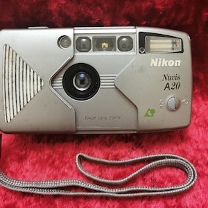 May include: A silver Nikon Nuvis A20 point-and-shoot camera with a black lens and a green arrow on the side. The camera has a strap attached.