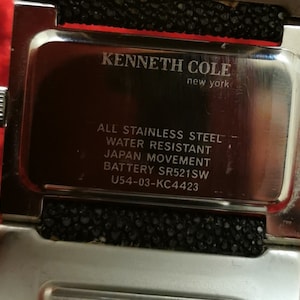 May include: A silver Kenneth Cole New York watch with a black band. The watch face is silver and has the text "ALL STAINLESS STEEL", "WATER RESISTANT", "JAPAN MOVEMENT", "BATTERY SR521SW", and "U54-03-KC4423" printed on it.
