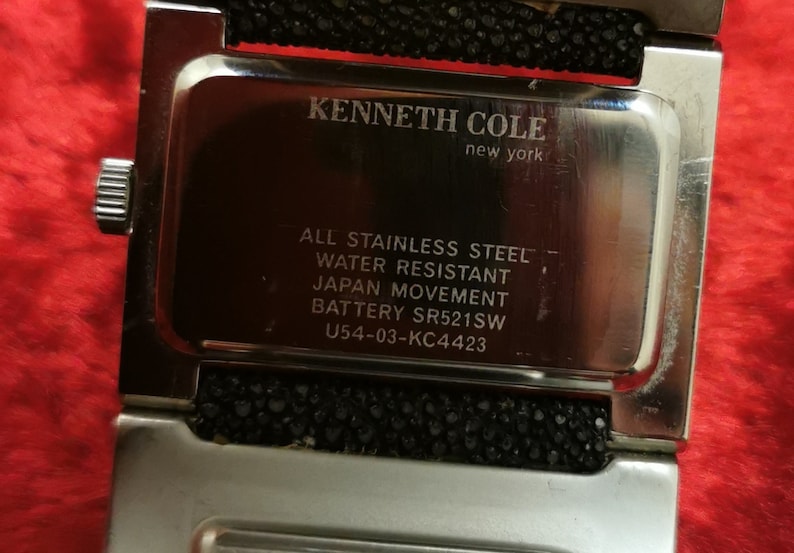 May include: A silver Kenneth Cole New York watch with a black band. The watch face is silver and has the text "ALL STAINLESS STEEL", "WATER RESISTANT", "JAPAN MOVEMENT", "BATTERY SR521SW", and "U54-03-KC4423" printed on it.