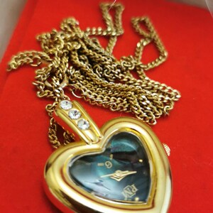 Op de afbeelding: Een goudkleurig hartvormig hangerhorloge met bijpassende ketting. De wijzerplaat van het horloge is donkergroenblauw met gouden wijzers en uurmarkeringen. Drie heldere kristallen sieren de bovenkant van het hart. Het horloge ligt op een rode ondergrond.