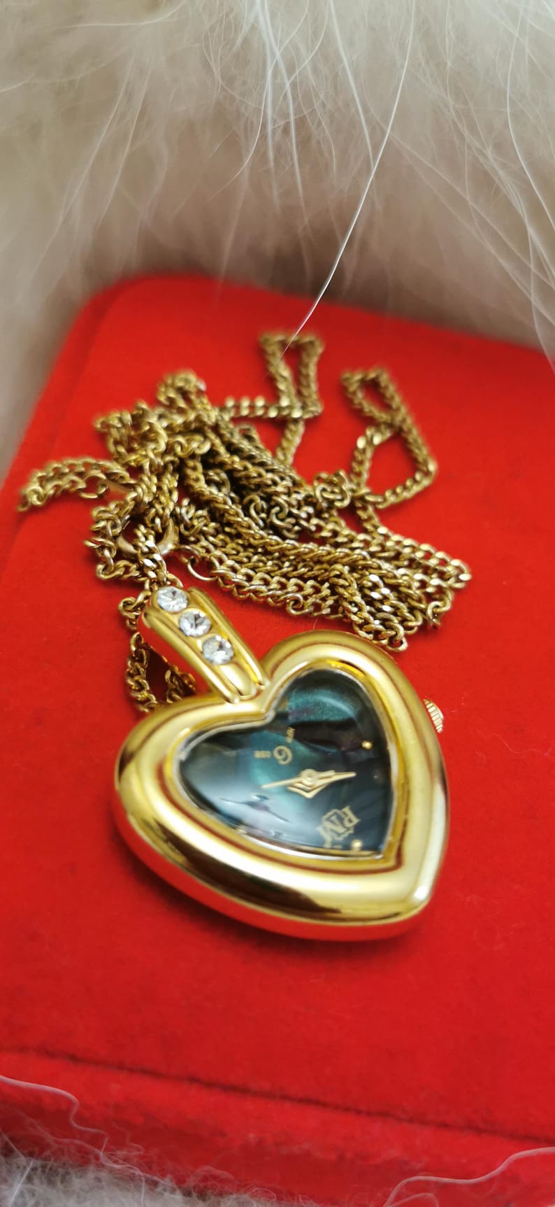 Op de afbeelding: Een goudkleurig hartvormig hangerhorloge met bijpassende ketting. De wijzerplaat van het horloge is donkergroenblauw met gouden wijzers en uurmarkeringen. Drie heldere kristallen sieren de bovenkant van het hart. Het horloge ligt op een rode ondergrond.