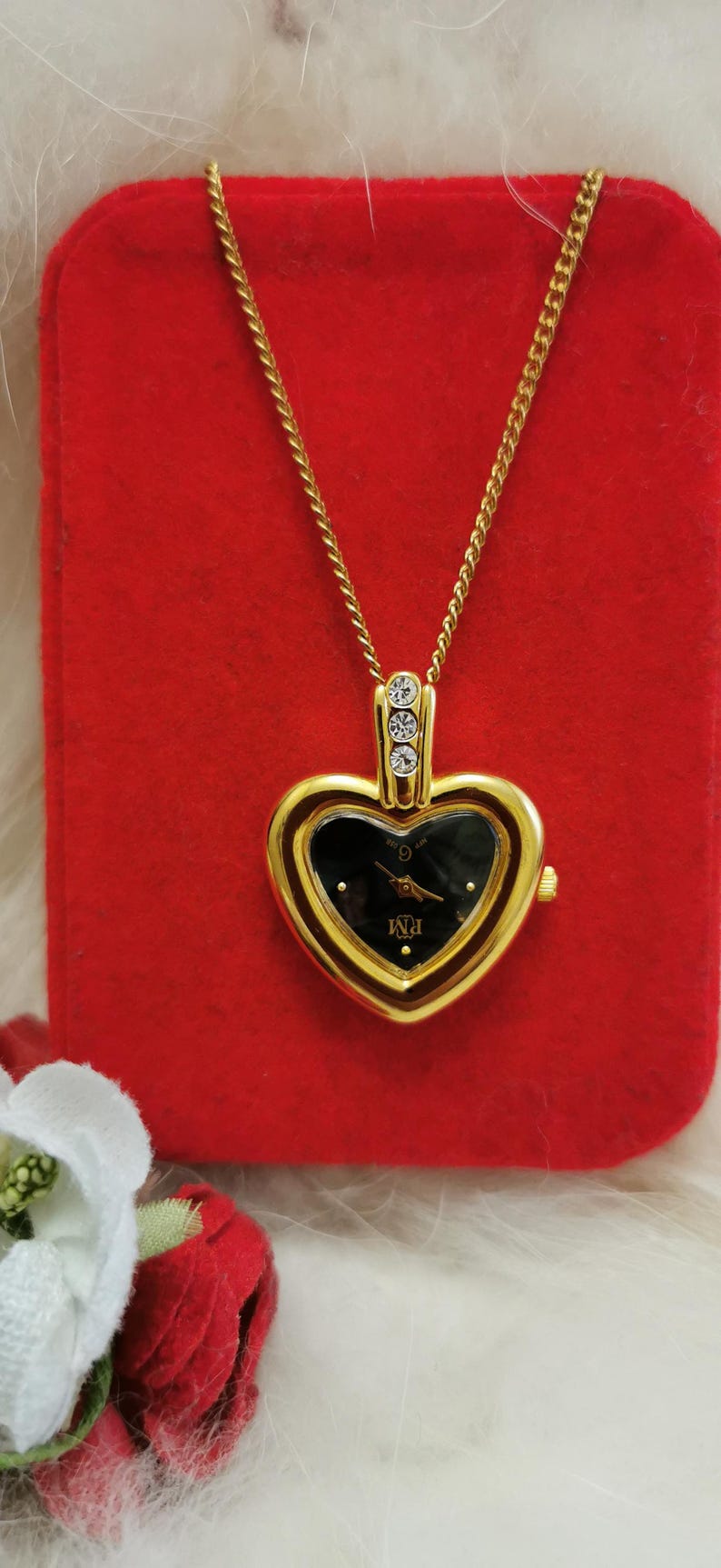 Op de afbeelding: Goudkleurig hartvormig hangerhorloge met een zwarte wijzerplaat en gouden wijzers, hangend aan een fijne gouden ketting. Het horloge heeft drie kleine, heldere kristallen aan de bovenkant. Het horloge wordt gepresenteerd op een rode fluwelen ondergrond.