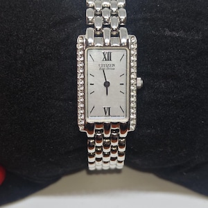 Könnte beinhalten: Eine silberne Citizen Eco-Drive-Uhr mit rechteckigem Zifferblatt und silbernem Armband. Das Zifferblatt hat einen hellen Hintergrund mit schwarzen Zeigern und römischen Ziffern. Die Uhr ist mit kleinen, klaren Kristallen verziert.