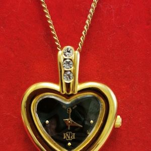 Op de afbeelding: Goudkleurige hartvormige hangerhorloge met een zwarte wijzerplaat en gouden wijzers. Het horloge heeft een gouden ketting en is versierd met drie kleine, heldere kristallen. De wijzerplaat toont de letters "PM".