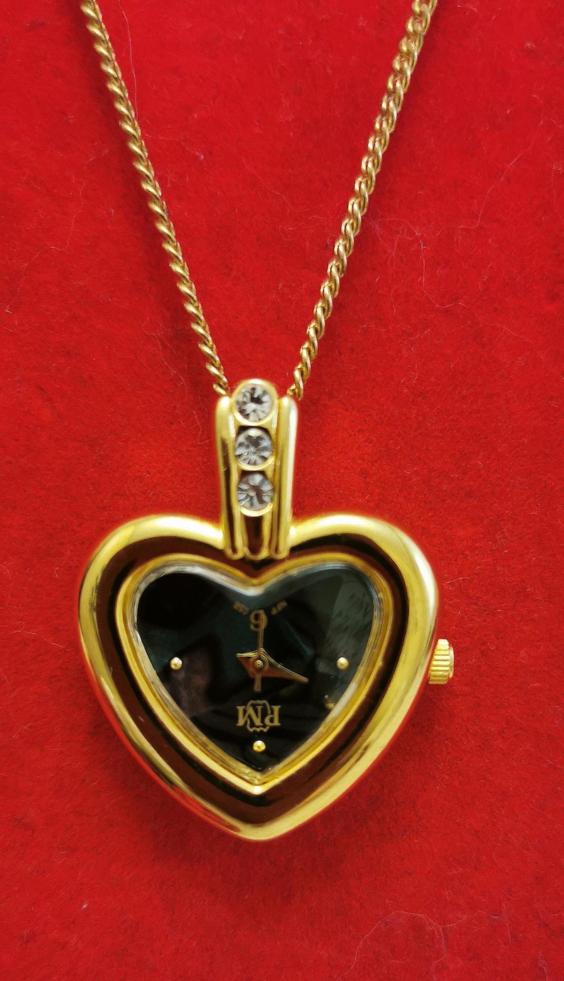 Op de afbeelding: Goudkleurige hartvormige hangerhorloge met een zwarte wijzerplaat en gouden wijzers. Het horloge heeft een gouden ketting en is versierd met drie kleine, heldere kristallen. De wijzerplaat toont de letters "PM".