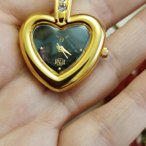 Op de afbeelding: Goudkleurig hartvormig hangerhorloge met een donkere wijzerplaat en gouden wijzers. Het horloge heeft een gouden ketting en drie heldere stenen aan de bovenkant. De wijzerplaat toont de letters "RM".