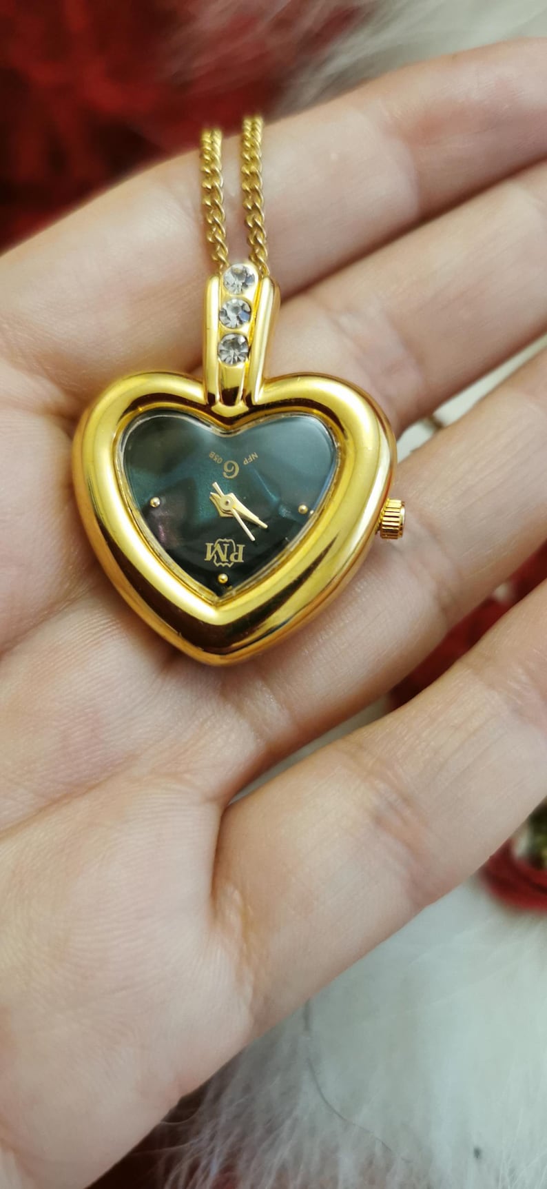 Op de afbeelding: Goudkleurig hartvormig hangerhorloge met een donkere wijzerplaat en gouden wijzers. Het horloge heeft een gouden ketting en drie heldere stenen aan de bovenkant. De wijzerplaat toont de letters "RM".