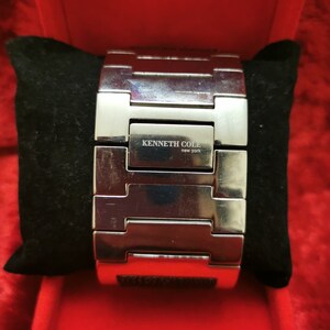 May include: A silver metal watch band with a black rectangular face. The watch band has a clasp and the face has the text "KENNETH COLE new york" on it.