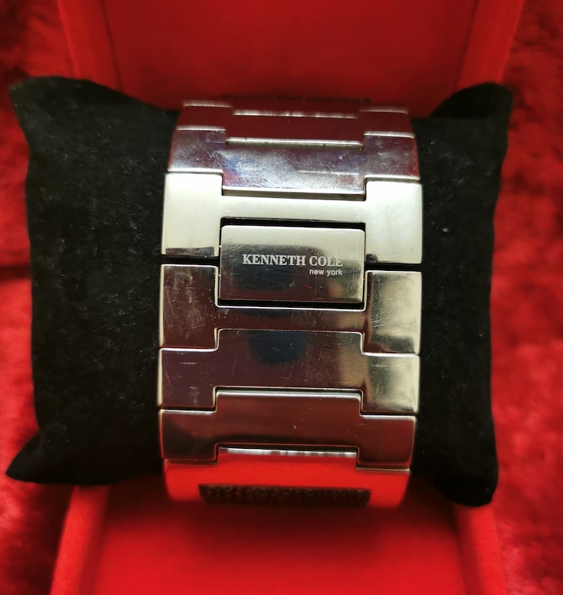 May include: A silver metal watch band with a black rectangular face. The watch band has a clasp and the face has the text "KENNETH COLE new york" on it.