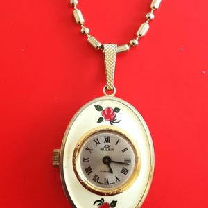 Könnte beinhalten: Eine goldene Kettenkette mit einem weißen ovalen Anhänger, der ein goldfarbenes Zifferblatt mit schwarzen römischen Ziffern und Zeigern zeigt. Das Zifferblatt trägt den Text "BULER 17 JEWELS" und ist mit roten Rosen verziert.