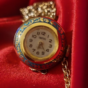 Puede incluir: Un reloj de bolsillo Smiths vintage con caja y cadena doradas. La esfera del reloj es blanca con números y manecillas negras. La caja está decorada con esmalte azul y dorado. El reloj está sobre un fondo de terciopelo rojo.