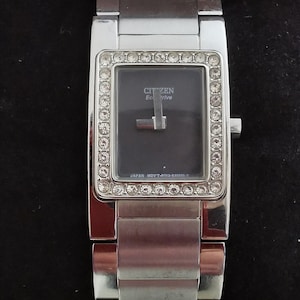 May include: A silver Citizen Eco-Drive watch with a rectangular face and a silver band. The face is surrounded by small clear crystals. The watch face is black and has the text "CITIZEN Eco-Drive" and "JAPAN MVT-B093-5045655 Y".