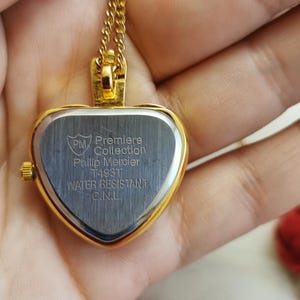 Op de afbeelding: Een goudkleurige hartvormige hangerhorloge met bijpassende ketting. De wijzerplaat van het horloge is zilverkleurig met de tekst "Premiere Collection Phillip Mercier" en "Water Resistant".