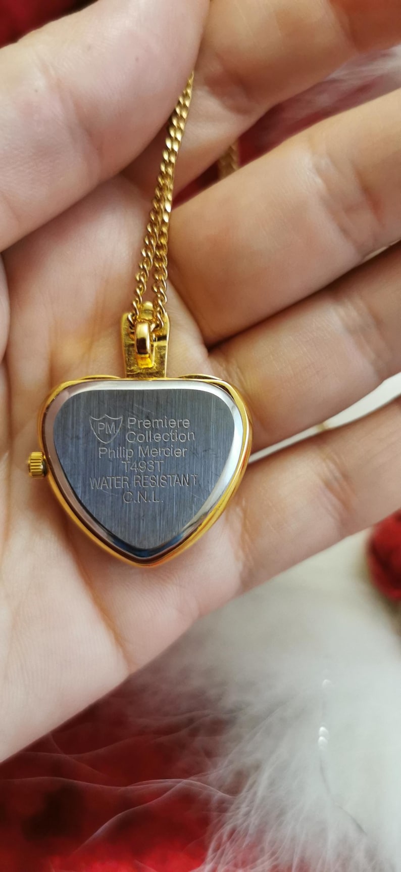 Op de afbeelding: Een goudkleurige hartvormige hangerhorloge met bijpassende ketting. De wijzerplaat van het horloge is zilverkleurig met de tekst "Premiere Collection Phillip Mercier" en "Water Resistant".