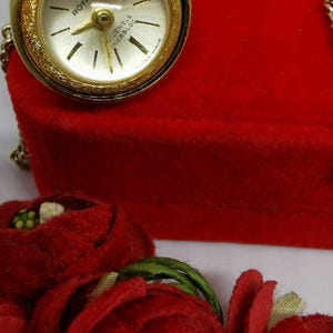 Pode incluir: Um relógio de bolso Rotary dourado com um mostrador branco e marcadores de hora romanos pretos. O relógio está em uma corrente dourada e está em uma caixa de veludo vermelho. O relógio é cercado por flores de veludo vermelho.