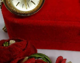 Może przedstawiać: Złoty zegarek kieszonkowy Rotary z białą tarczą i czarnymi rzymskimi cyframi godzinowymi. Zegarek jest na złotym łańcuszku i znajduje się w czerwonym pudełku z aksamitu. Zegarek jest otoczony czerwonymi aksamitnymi kwiatami.