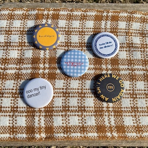 Op de afbeelding: Een verzameling van vijf ronde buttons met verschillende ontwerpen en tekst. Eén button heeft de tekst "the you all day ng" op een gele achtergrond. Een andere zegt "ooo my tiny dancer!". Andere buttons zeggen "baby I'll do the dishes!", "my boyfriend is on stage!!!", en "HERE WE GO AGAIN".