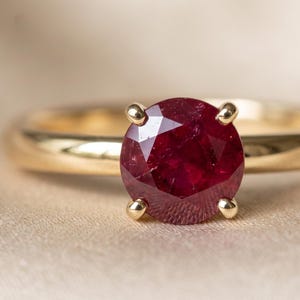May include: A gold ring featuring a round, deep red ruby gemstone set in a four-prong setting. The band is smooth, polished, and circular, showcasing a classic design with a vibrant, faceted centre stone that catches the light.
