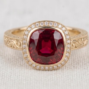 Peut inclure: Une bague en or avec une grande pierre précieuse rouge foncé taille coussin au centre, entourée d'un halo de petites pierres blanches. L'anneau en or est orné de motifs complexes en relief sur les côtés de la tige de la bague.