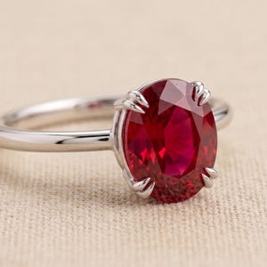 May include: A silver-toned ring featuring a large, oval-cut, deep red gemstone set in a six-prong mounting. The band is smooth and polished, showcasing a classic design that highlights the vibrant color and faceted surface of the central stone.