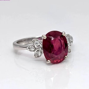 May include: A ring with a large oval ruby gemstone surrounded by smaller diamonds set in a white gold band. The diamonds are set in a leaf design.
