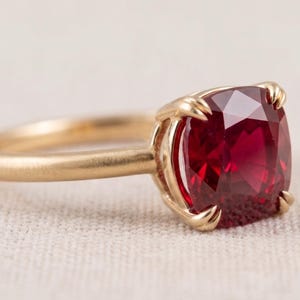 May include: A gold ring featuring a cushion-cut deep red gemstone set in a four-prong setting. The band has a smooth, polished finish and a rounded profile. The vibrant red stone is faceted, reflecting light across its surface within the gold metal.