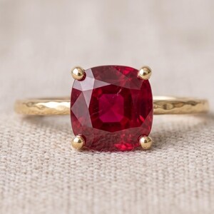 Puede incluir: Un anillo de oro con una gran piedra preciosa de color rojo intenso en talla cojín, sujeta por cuatro garras. La banda de metal tiene un acabado martillado texturizado, lo que proporciona un patrón de superficie que contrasta con la piedra.