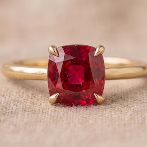 May include: A gold ring featuring a cushion-cut red gemstone set in a four-prong setting. The band is smooth and polished, showcasing a simple design that highlights the vibrant color and faceted surface of the central stone.