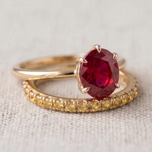 May include: A gold ring featuring a large oval-cut deep red ruby held by six prongs, stacked with a second gold band encrusted with a continuous row of small, round-cut yellow gemstones that encircle the entire circumference of the band.