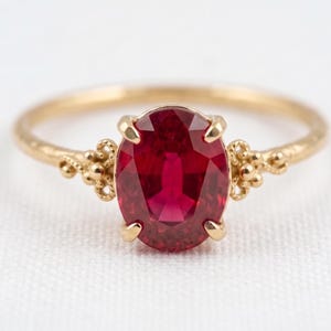 May include: A gold ring featuring a central oval-cut deep red gemstone held by four prongs. The band has a textured finish and decorative metalwork details on both sides of the stone, creating a detailed and ornate appearance for this piece of jewelry.