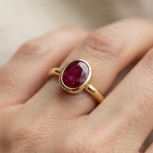 Op de afbeelding: Een gouden ring met een centrale ovaalvormige dieprode edelsteen in een gladde zetting. De ringband is van gepolijst geelgoud met een eenvoudig, afgerond profiel, wat de levendige kleur en het gefacetteerde oppervlak van de steen benadrukt.