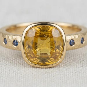 May include: A gold ring featuring a large, cushion-cut yellow gemstone set in a smooth bezel. The band has a hammered texture and is accented with small, round blue gemstones embedded along the sides of the shank, creating a contrast.