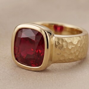 May include: A gold ring featuring a large, square-cut, deep red gemstone set in a smooth bezel. The band of the ring has a textured, hammered finish, and a small, heart-shaped red accent is visible on the inner surface of the gold band.
