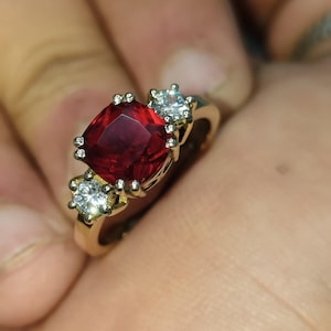 May include: A gold ring with a large, red, square-shaped gemstone surrounded by smaller, clear gemstones.