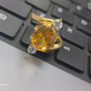 Puede incluir: Un anillo de oro con una gran piedra preciosa ovalada de color ámbar y dos piedras preciosas más pequeñas de color claro. El anillo está sobre un teclado negro.