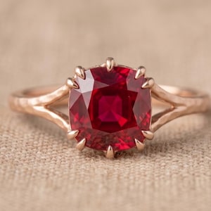 May include: A rose gold ring featuring a cushion-cut deep red ruby gemstone held by eight prongs. The band has a split shank design with a textured, hammered finish, creating a distinct look for this piece of fine jewelry.