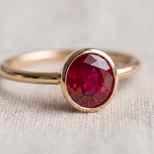 May include: A gold ring featuring a single oval-cut deep red ruby gemstone set in a smooth bezel setting. The band is thin and polished, showcasing a metallic yellow gold finish that complements the vibrant, faceted red stone at the center.